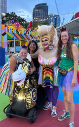 Group photo at a colorful downtown outdoor festival: drag performer in a gold headdress and rainbow costume poses with three friends, one on a sequined unicorn scooter, with city skyline and string lights in the background.