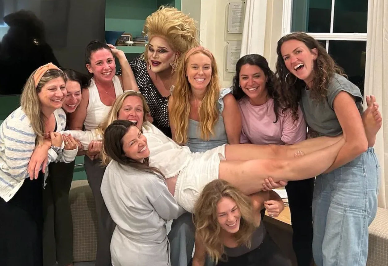 Group of smiling women at an indoor home gathering lift a laughing friend horizontally, posing with a glam drag performer in a big wig and sequined outfit.