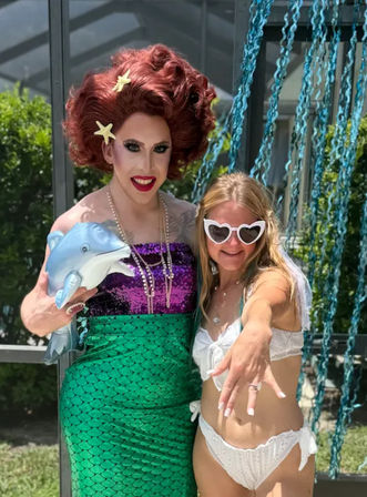 Drag performer in red wig and mermaid sequin dress holding an inflatable dolphin poses with a woman in a white bikini and heart-shaped sunglasses at a sunny backyard poolside party with blue streamers.