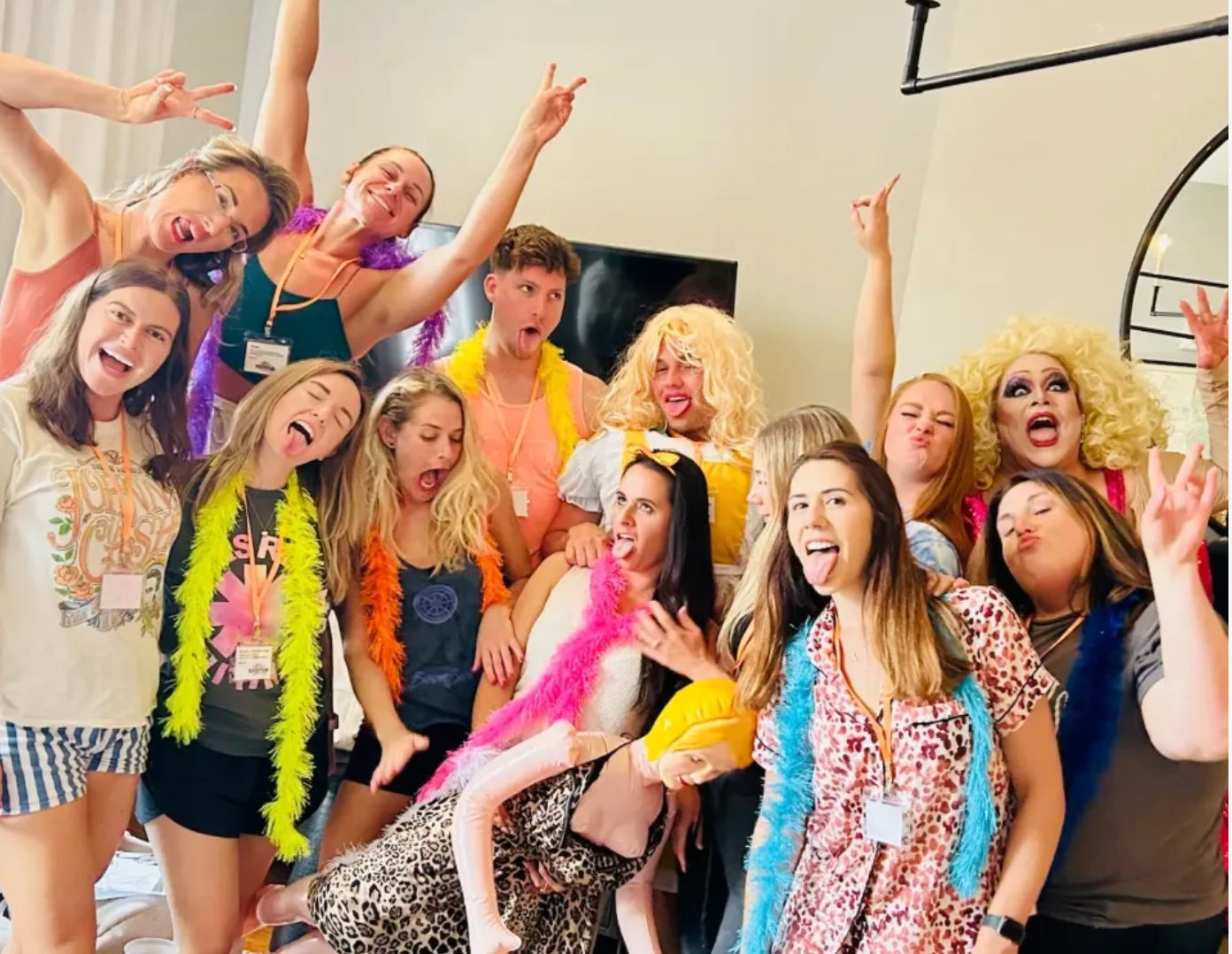 Energetic indoor group photo of friends in a bright living room wearing colorful feather boas and playful costumes, striking silly poses with tongues out and hands raised, featuring a drag performer in a big blonde wig and an inflatable doll.
