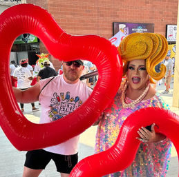 Two people at an outdoor Pride festival holding oversized red inflatable hearts — a drag performer in a sparkling sequin dress, pearl necklace and large yellow wig smiling, and a person in sunglasses and a graphic t-shirt posing behind the heart with a brick building and crowd in the background.