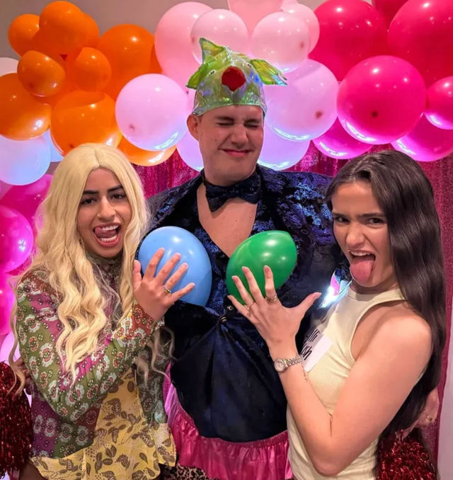 Playful costume party photo booth with three people in front of a pink-orange balloon arch — a costumed person holds blue and green balloons at their chest while two friends make silly faces and stick out their tongues.