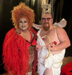 Two costumed performers smiling against a dark backdrop: a drag artist in a voluminous orange wig, red feathered jacket and leopard-print outfit, alongside a playful cupid in a white toga with wings, heart-shaped sunglasses and a gold bow with heart-tipped arrow.