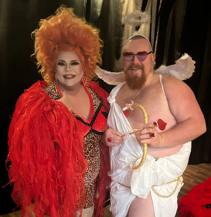 Two costumed performers smiling against a dark backdrop: a drag artist in a voluminous orange wig, red feathered jacket and leopard-print outfit, alongside a playful cupid in a white toga with wings, heart-shaped sunglasses and a gold bow with heart-tipped arrow.