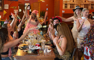 Lively bachelorette party in a restaurant: a drag performer in a pink feathered costume sings into a microphone while guests at a long table cheer, clap, and toss dollar bills amid drinks, pitchers, and plates.