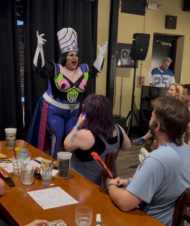 Drag performer in a colorful villain-style costume with tall white-and-purple headdress, neon green-and-pink chest design and blue cape, energetically entertaining a seated bar/cabaret audience as patrons photograph and cheer.