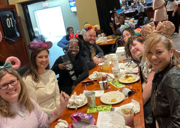 Smiling group of friends wearing colorful novelty mouse ears and headbands gathered around a long wooden table for brunch in a busy casual restaurant, with plates, drinks and menus visible.