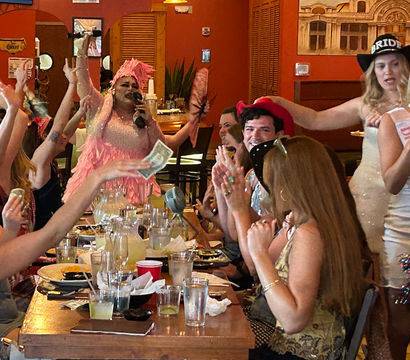 Lively bachelorette party in a restaurant with a drag performer in a pink feathered costume singing at a long table while guests cheer, toss dollar bills, and a woman wearing a bride-to-be sash smiles.
