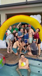 Backyard pool party group photo under a large inflatable rainbow arch, featuring a sparkly drag performer, friends with drinks, and a man hugging a beach ball in the pool.