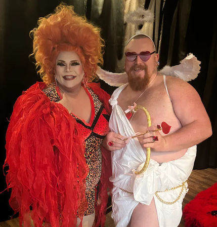 Two performers posing indoors: a drag queen in leopard-print bodysuit, red feathered coat and towering orange wig, and a bearded Cupid in a white toga with wings, heart-shaped sunglasses and a gold bow with heart-tipped arrow.
