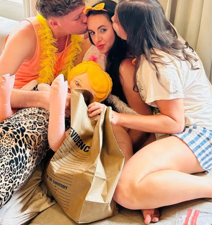 Three friends lounging on a living-room couch at a playful party — two kiss the woman in the center who holds a blond inflatable doll and a large 'EVIDENCE' paper bag, with a yellow feather boa and leopard-print leggings adding color.