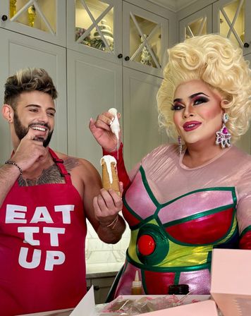 Two people in a bright kitchen decorating a pastry cone—one wearing a red “EAT IT UP” apron smiling, the other in glamorous drag with a big blonde wig and colorful costume piping white frosting onto the dessert.