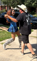 Drag performer in a blue sequined costume and silver knee-high boots dancing with a man in a cowboy hat and black shirt on a suburban driveway