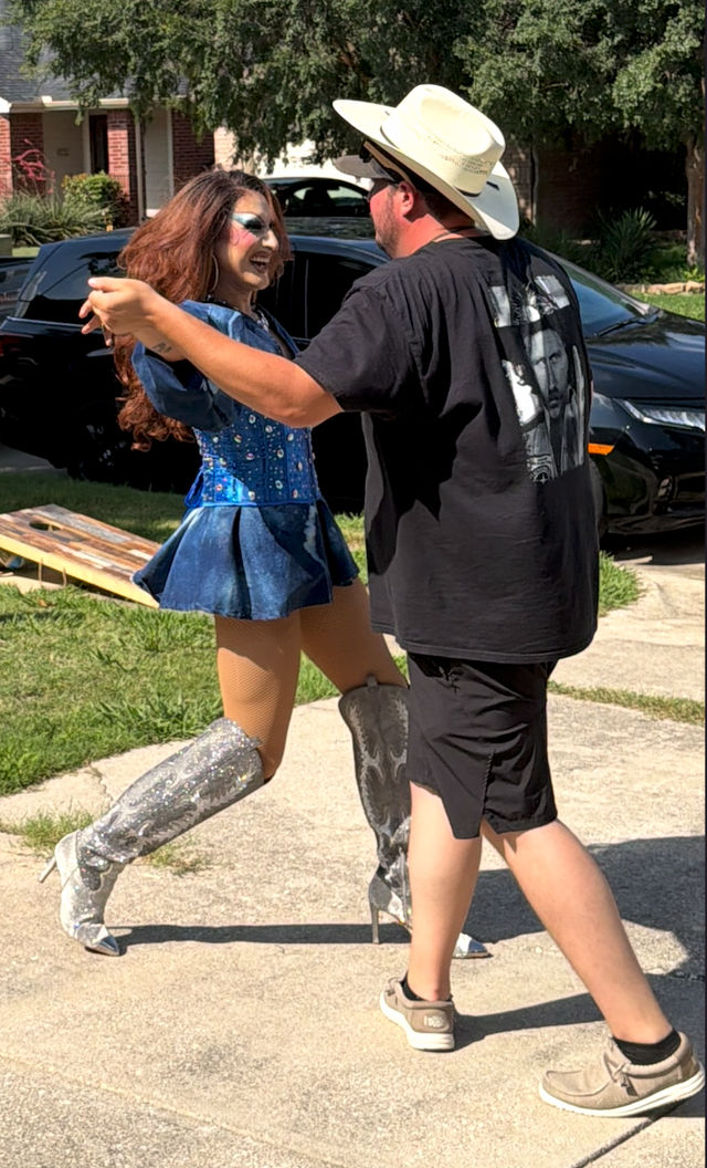 Drag performer in a blue sequined costume and silver knee-high boots dancing with a man in a cowboy hat and black shirt on a suburban driveway