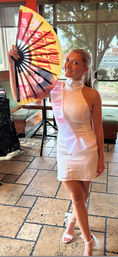 Smiling woman in a white dress and 'Bride to Be' sash holding a large colorful 'All You Can Eat' fan in a sunlit restaurant with tiled floor and big windows.