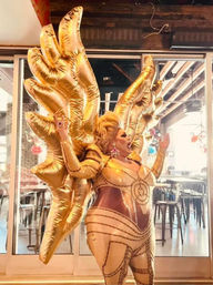 Drag performer in a shimmering gold bodysuit and oversized inflatable gold wings striking a dramatic pose inside an urban bar or restaurant with glass doors.