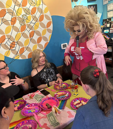 Drag performer with big blonde wig and pink outfit holding a microphone entertaining guests at an indoor rainbow-themed cookie decorating party with colorful plates, sprinkles and dollar tips