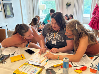 Three women leaning over a cluttered table in a bright living room, pointing at papers and collaborating on paperwork during a casual workshop; phones, craft supplies, a drink can and lanyard name badges visible, others chatting on sofas in the background.