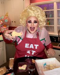 Person with voluminous blonde wig and dramatic makeup smiling in a colorful home kitchen, wearing a red apron that reads "EAT IT" while holding a decorated yellow cookie amid cookie-decorating supplies on the countertop.