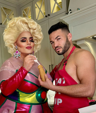 Drag performer in a blonde wig and colorful metallic dress piping cream into a novelty pastry as a man in a red apron holds it in a modern home kitchen
