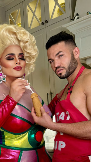 Drag performer in a blonde wig and colorful metallic dress piping cream into a novelty pastry as a man in a red apron holds it in a modern home kitchen