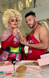 Glamorous drag performer in a platinum wig and metallic colorful dress and a shirtless man in a red apron decorating novelty cookies in a bright home kitchen, with cookie boxes and decorating supplies on the marble counter.