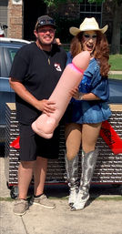 Smiling man and costumed performer in a suburban driveway posing with a large pink inflatable novelty prop by a birthday-wrapped cart; performer wears a cowboy hat, blue outfit and silver sequined boots.