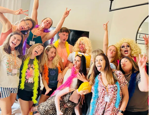 Playful group of adults in a living room wearing bright feather boas and wigs, striking energetic poses and making silly faces around an inflatable prop for a fun party photo.
