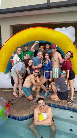 Backyard pool party group posing under a large inflatable rainbow arch, friends in swimsuits and casual wear, one person in a blue sequined drag outfit, man hugging a beach ball in the pool.