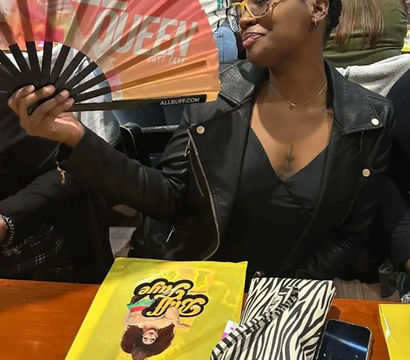 Young woman in a black leather jacket and yellow glasses playfully fanning herself with a colorful hand fan reading 'SIZE QUEEN' while seated at a busy indoor restaurant table with a zebra-print clutch, smartphone and bright yellow menu.
