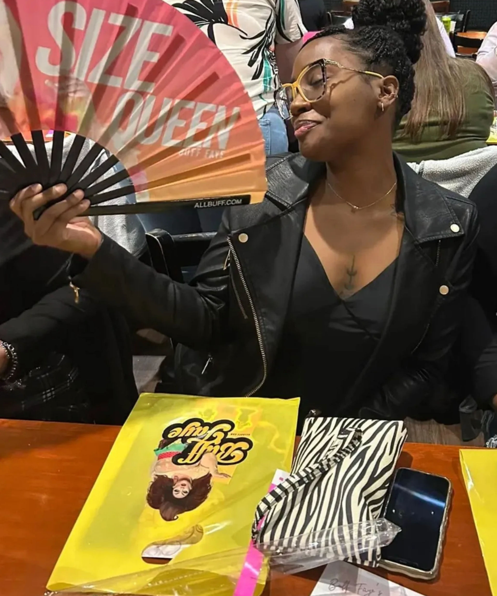 Young woman in a black leather jacket and yellow glasses playfully fanning herself with a colorful hand fan reading 'SIZE QUEEN' while seated at a busy indoor restaurant table with a zebra-print clutch, smartphone and bright yellow menu.