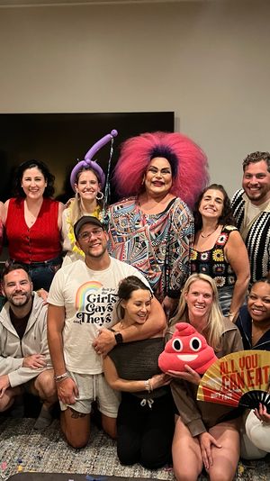 Lively living-room group photo of friends at an indoor party, centered on a drag performer with a large pink wig and colorful patterned dress, with people smiling, kneeling and holding a red plush toy and decorative fan, casual summer outfits.