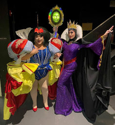 Backstage at a theater, two performers in vibrant fairy-tale costumes: a Snow White–inspired dress with oversized puffed sleeves and a red bow, and a glittering purple Evil Queen with crown holding a glowing green mirror.