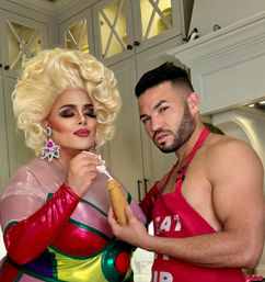 Glam drag queen with voluminous blonde wig and colorful costume pipes frosting onto a pastry held by a shirtless man in a red apron in a bright home kitchen.