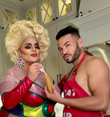 Glam drag queen with voluminous blonde wig and colorful costume pipes frosting onto a pastry held by a shirtless man in a red apron in a bright home kitchen.