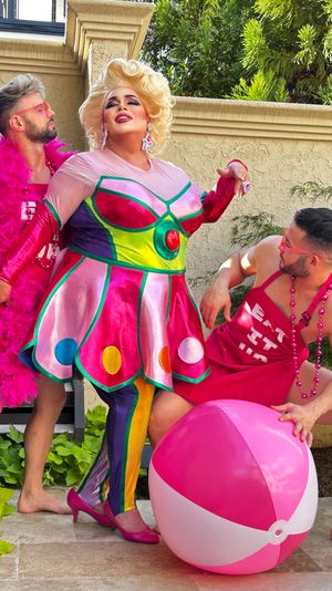 Drag performer in a colorful metallic retro dress and blonde wig poses on a sunny backyard patio with two men in pink outfits, a large pink-and-white beach ball, stucco wall and greenery