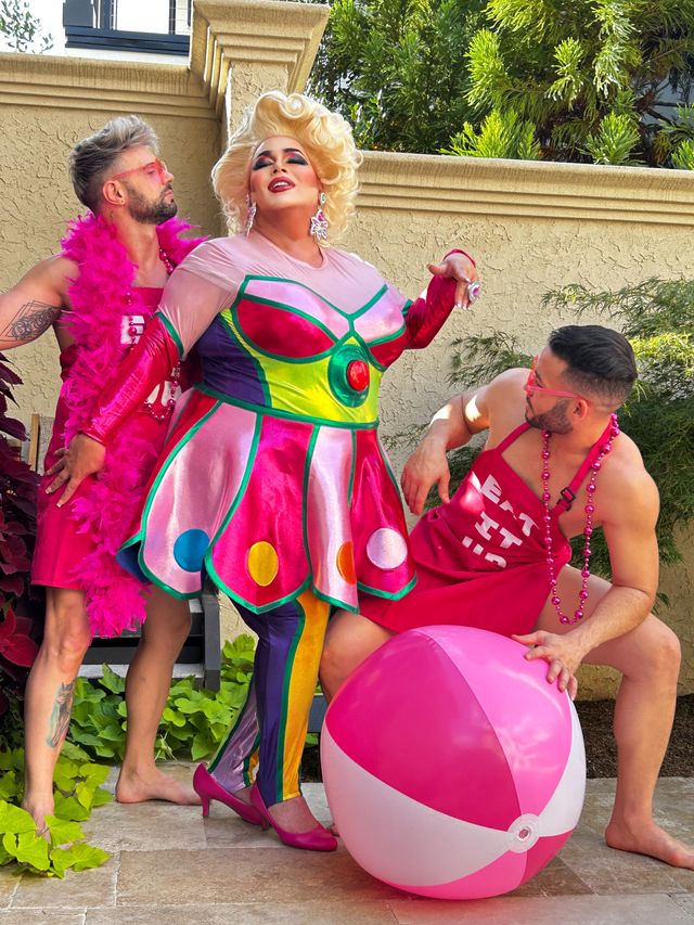 Drag performer in a colorful metallic retro dress and blonde wig poses on a sunny backyard patio with two men in pink outfits, a large pink-and-white beach ball, stucco wall and greenery