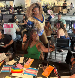 Energetic drag performer in a gold-and-blue sequined costume dances through a lively indoor bar, waving cash while patrons take selfies at tables with rainbow and trans pride flags.