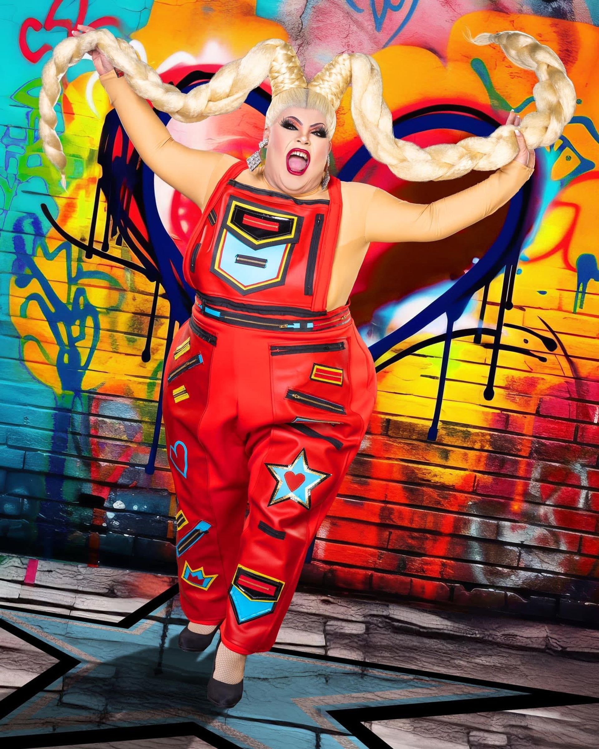 Drag performer in red patched overalls and dramatic makeup, swinging giant blonde braided pigtails like horns in front of a colorful heart graffiti mural on a brick urban street-art backdrop.