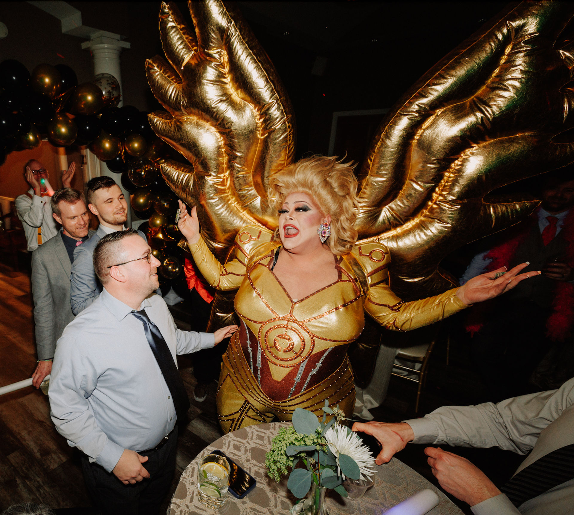 Drag performer in a shimmering gold winged costume flamboyantly entertaining smiling guests at an indoor party with black-and-gold balloons and drinks on a cocktail table.