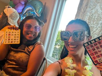 Two people at an indoor bingo night, wearing oversized jeweled sunglasses and leis, smiling and holding bingo cards amid colorful balloons and a dolphin foil balloon by a sunny window.