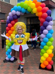 Drag performer in a SpongeBob-themed outfit and rainbow socks posing under a vibrant rainbow balloon arch at an outdoor Pride celebration entrance