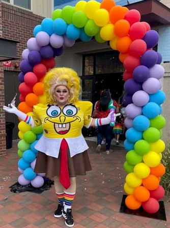 Drag performer in a SpongeBob-themed outfit and rainbow socks posing under a vibrant rainbow balloon arch at an outdoor Pride celebration entrance