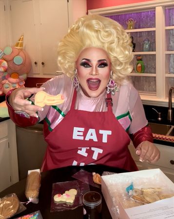 Drag performer with big blonde wig and bold makeup in a bright home kitchen, wearing a red apron printed “EAT IT” and holding a decorated cookie amid baking supplies