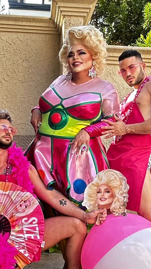 Three people posing on a sunny outdoor patio: a smiling drag performer in a shiny pink-green costume and blonde wig flanked by two men in bold pink outfits — one with a feather boa and fan, the other holding a pink-and-white beach ball, playful summer party vibe.