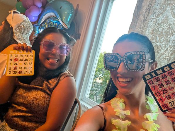 Two friends at a home bingo party wearing glittery oversized sunglasses and leis, smiling and holding up bingo cards with colorful balloons (including a dolphin foil) and a sunlit window behind them.