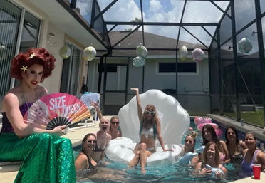 Backyard screened-patio pool party with friends gathered around a white inflatable seashell float and a drag performer in a green mermaid skirt holding a colorful fan