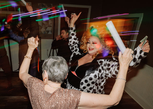 Drag performer in a polka-dot coat and big blonde wig dances on an indoor dance floor with a guest waving a foam light stick and dollar bill, colorful neon light streaks and lively party vibes.
