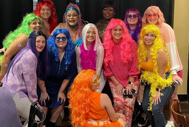 Festive group photo of people wearing bright colorful wigs and playful costumes (green, blue, pink, yellow, orange) smiling and posing at an indoor costume party.