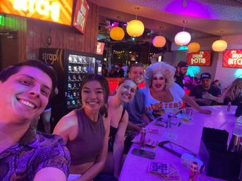 Group selfie with friends and a drag performer at a vibrant neon-lit bar during bingo night, colorful hanging lanterns, cocktails on the counter and lively city nightlife vibes.